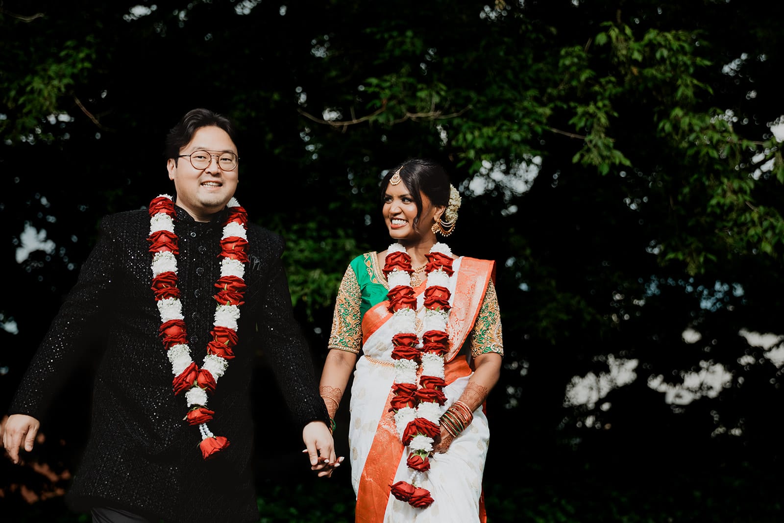 Un pas vers demain Couple de jeunes mariés d’origines indienne et coréenne souriants, main dans la main, lors d’un mariage multiculturel