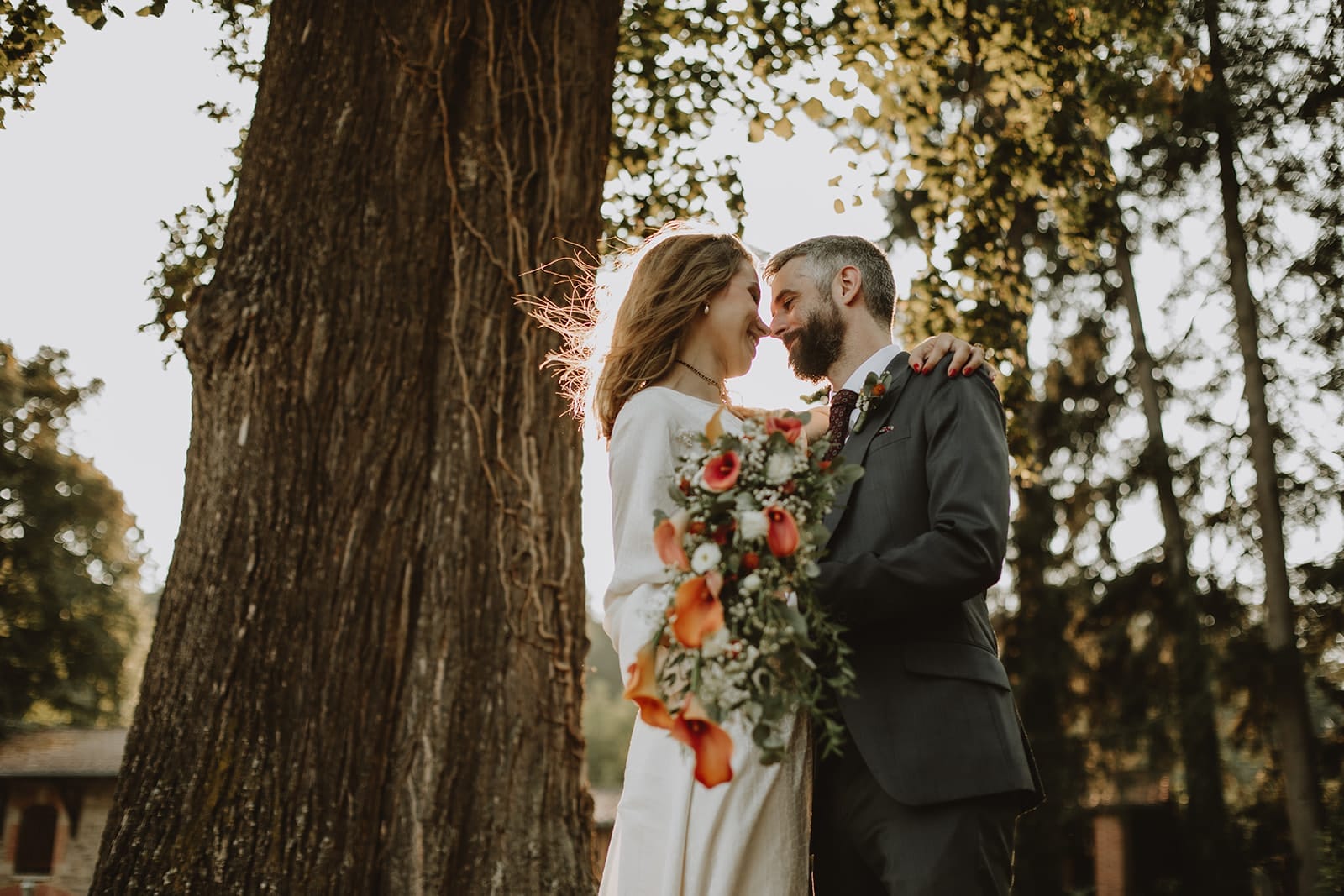 Couple de mariés au Manoir de Tourville enlacés au pied d’un arbre centenaire, bouquet coloré et lumière dorée en fin de journée.