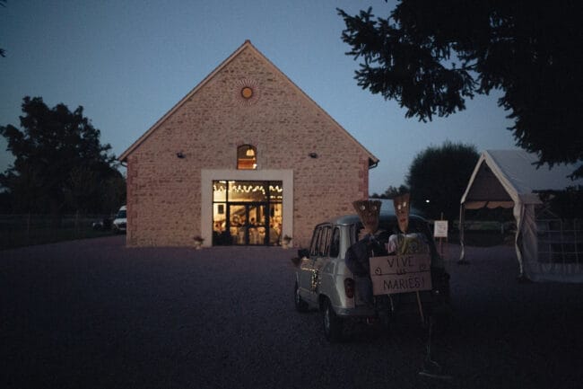 Voiture décorée avec pancarte “Vive les mariés” devant la salle du Domaine de la Pierre au crépuscule