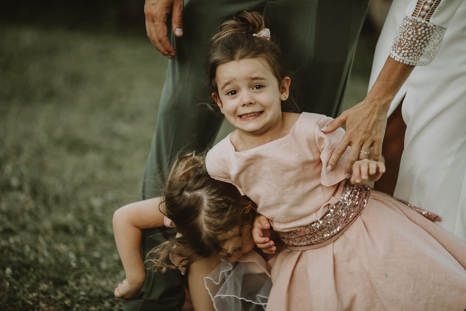 Deux petites filles en robe de cérémonie, l’une riant nerveusement, l’autre chutant, entourées d’adultes tentant de les retenir.