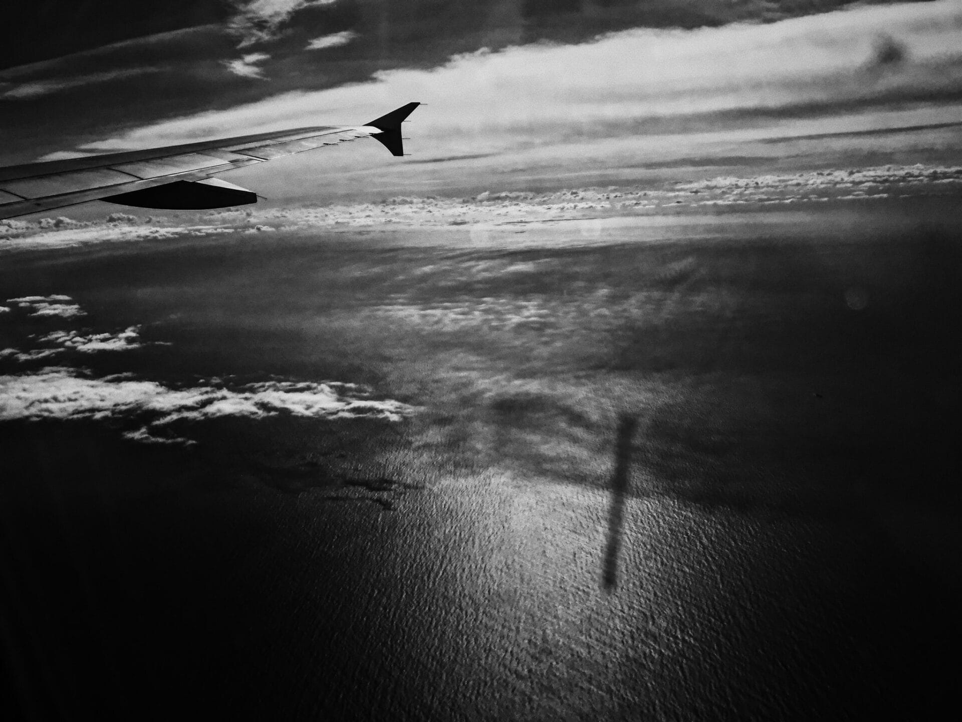 Vue aérienne en noir et blanc depuis un avion, l’aile survolant un océan baigné de lumière.
